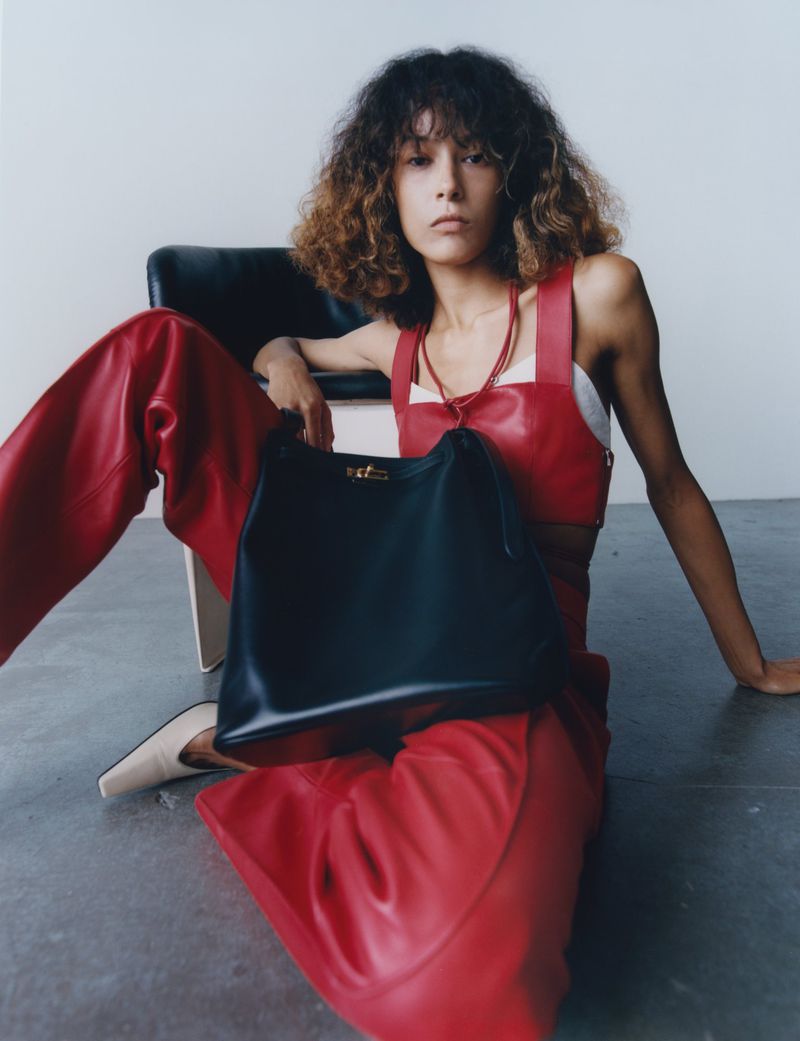 Model with curly hair wearing red leather trousers and top holding a black handbag in an interior setting
