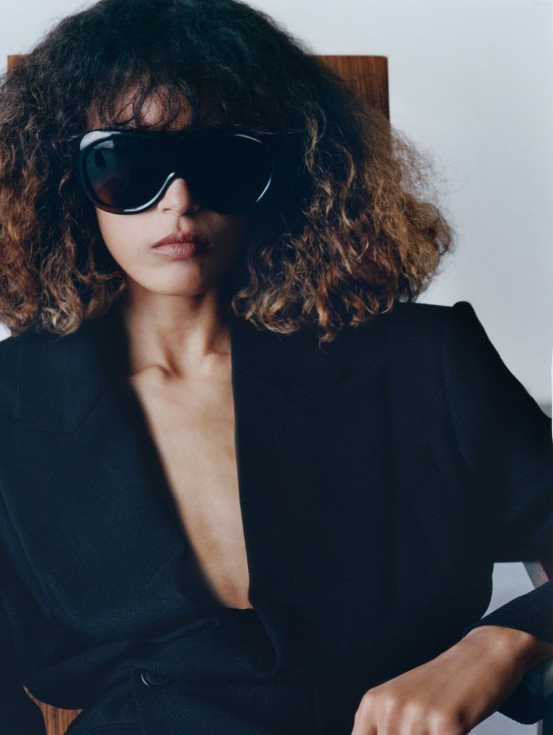 Portrait of a woman wearing sunglasses and a black jacket, sitting in a wooden chair in a studio setting with neutral background.