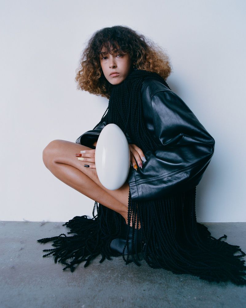 Model Prinnie Stott squatting in a black fringed leather jacket holding a white oval prop for Puss Puss Magazine
