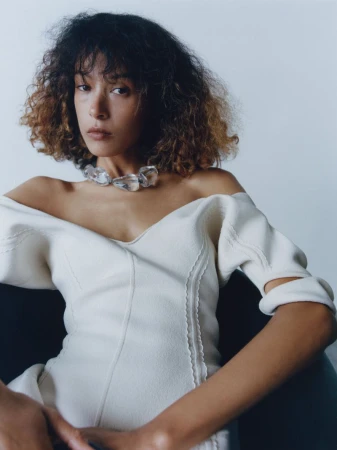 Model Prinnie Stott wearing a white off-shoulder dress and statement stone necklace against a plain background