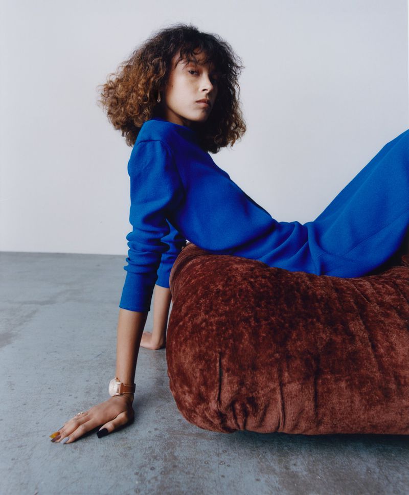 Model wearing blue top leaning against brown textured furniture in a studio setting