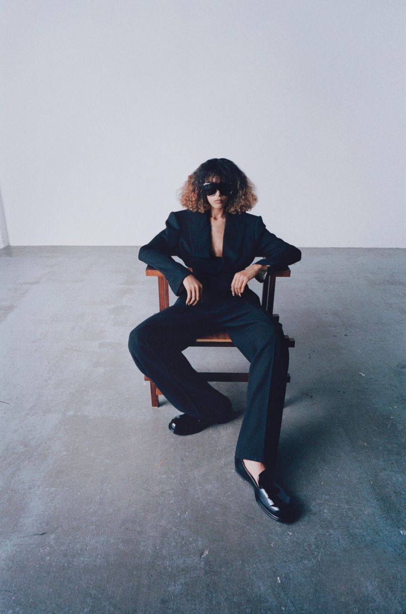 Model wearing a black suit and sunglasses seated on a wooden chair against a bare concrete wall