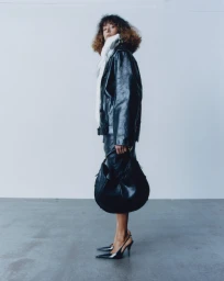 Model in a black leather jacket and matching skirt holding a black handbag against a light grey studio background.