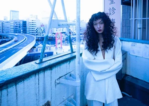 Model Suzune Oda with dark curly hair wearing a white jacket standing on a rooftop overlooking a city highway for 5ELEVEN Magazine