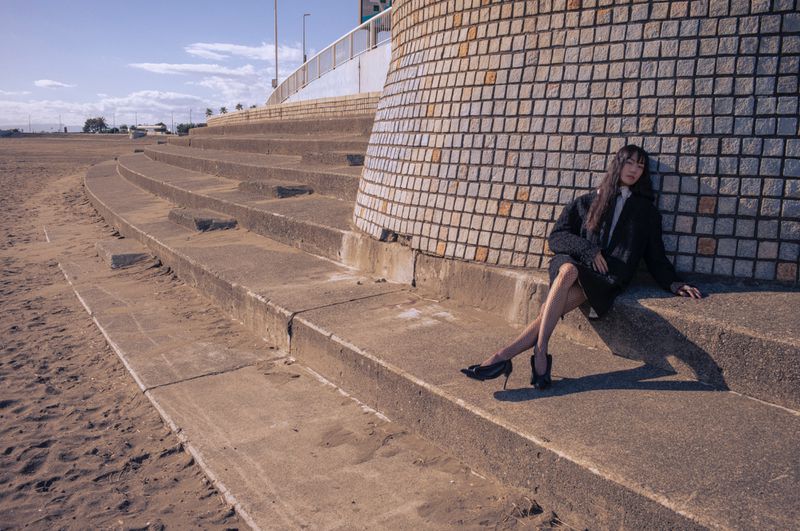 Model seated on concrete steps leaning against a tiled column near a sandy coastal area under a clear sky.