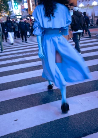 Model in a bright blue dress walking away across a pedestrian crosswalk surrounded by crowds in a city at dusk