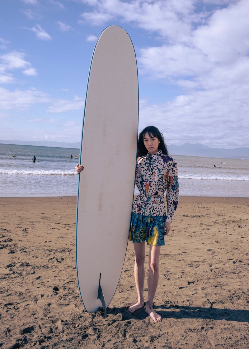 Model standing on a sandy beach holding a tall surfboard upright under a cloudy blue sky.