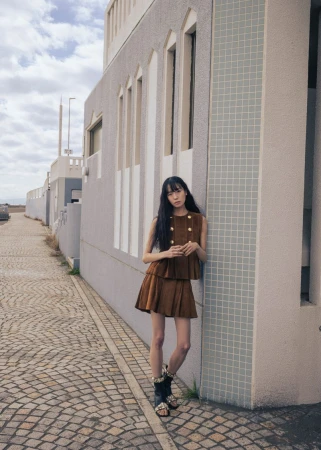 Model leaning against a pale textured building on a cobbled street, wearing a brown two-piece set.