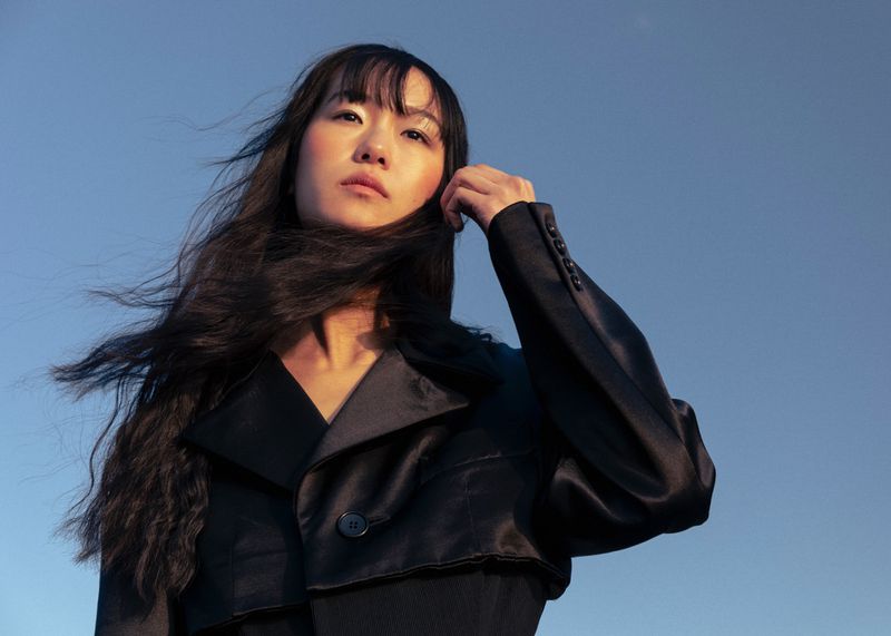 Model with long dark hair wearing a black jacket against a clear blue sky