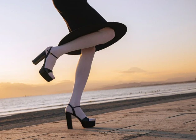 Model wearing white stockings and black platform heels holding a hat against a sunset coastal backdrop.