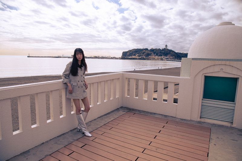 Woman in patterned jacket and white boots standing on a coastal terrace by the sea for 5ELEVEN Magazine