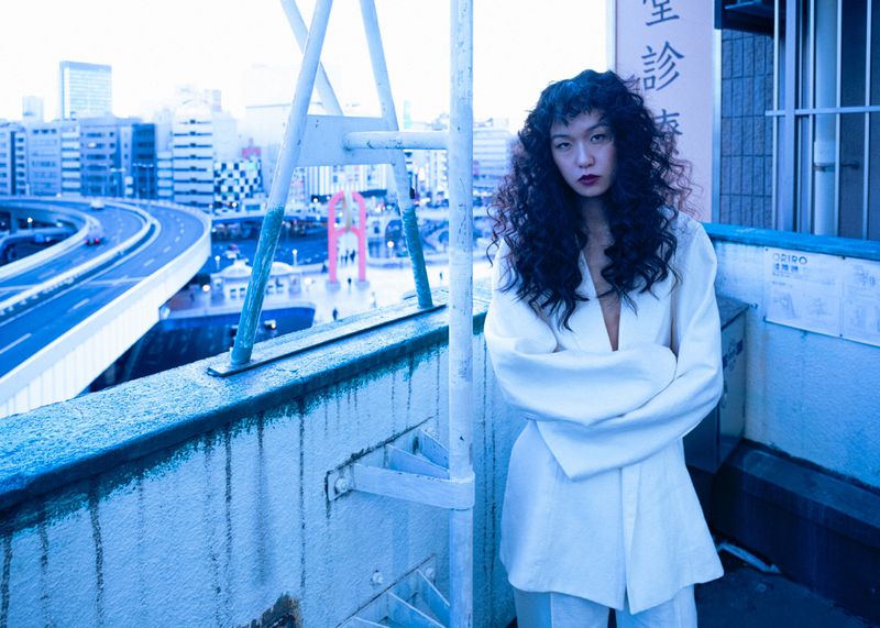 Model Suzune Oda with dark curly hair wearing a white jacket standing on a rooftop overlooking a city highway for 5ELEVEN Magazine