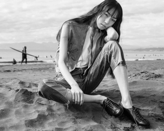 Black and white portrait of a model sitting on a sandy beach wearing a denim vest and dark shoes, with ocean waves in the background.