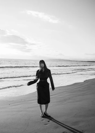 Model standing on a sandy beach wearing a dark belted coat in a monochrome fashion editorial photograph.