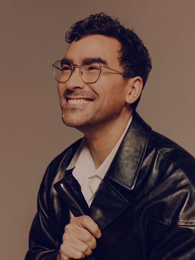 Smiling man in a black leather jacket and glasses looking off to the side in a studio shot with a neutral background.