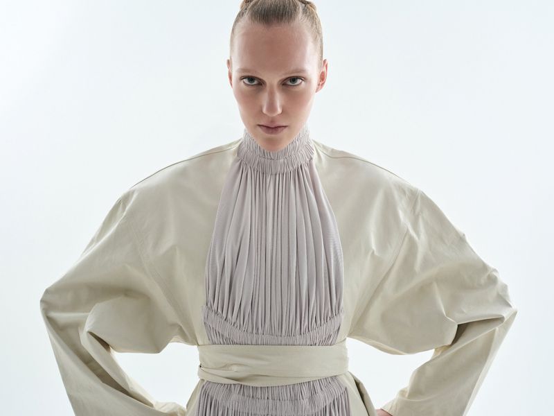 Model in a high-necked cream pleated dress with puffed sleeves against a solid white background