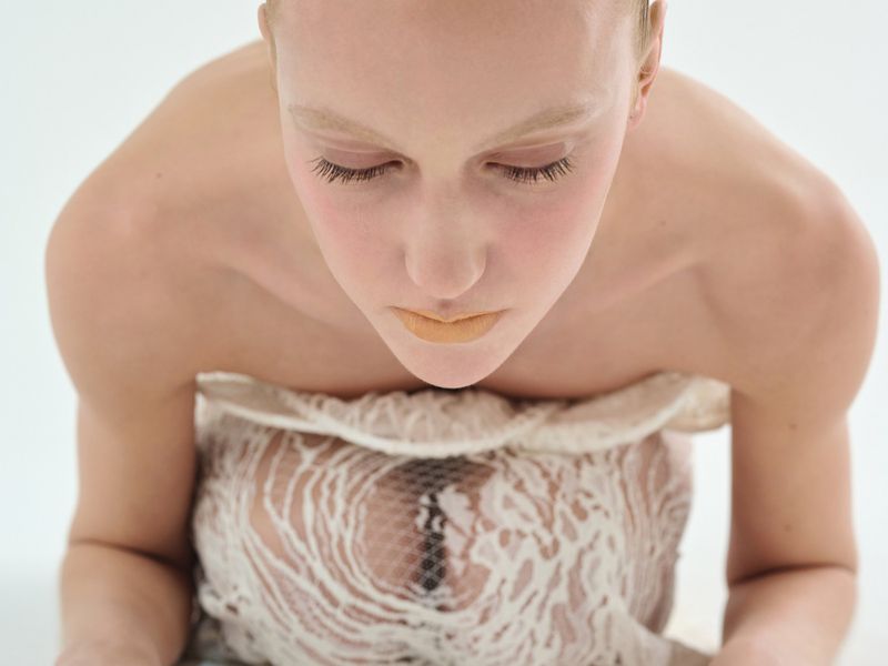Close-up high-angle portrait of a model with closed eyes wearing a textured cream lace top against a plain white background.