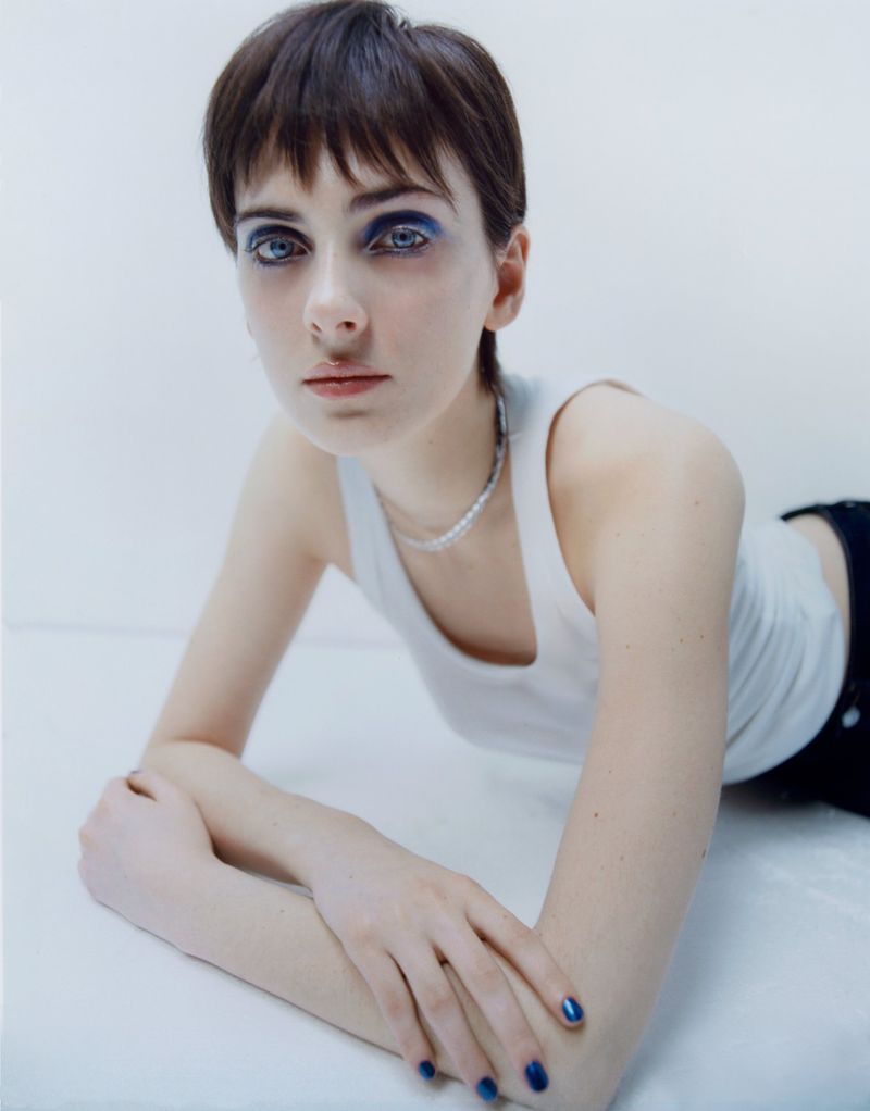 Model with blue eyeshadow and a white tank top leaning forward on a seamless white studio background