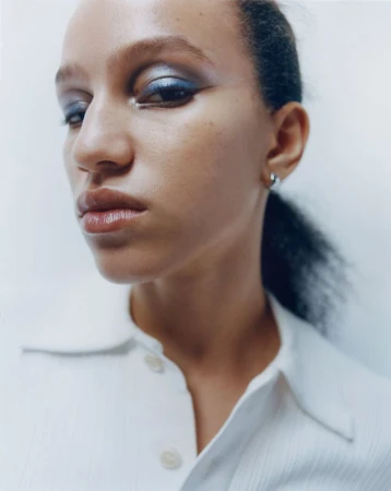 Close-up portrait of a woman wearing blue eye makeup and a white collared shirt against a white backdrop.