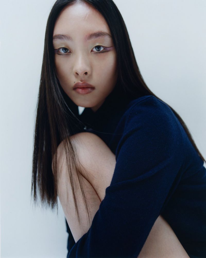 Asian woman with long black hair poses in a navy turtleneck sweater against a white background in a studio portrait with a shallow depth of field.