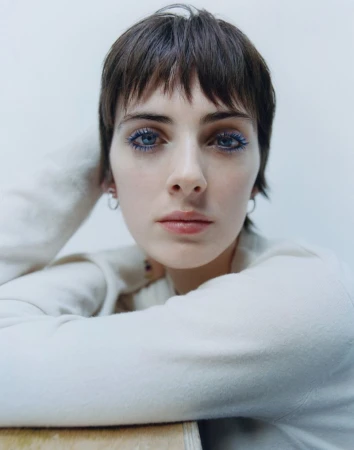 Close-up portrait of a woman with short dark hair and blue mascara wearing a white sweater against a seamless light background.