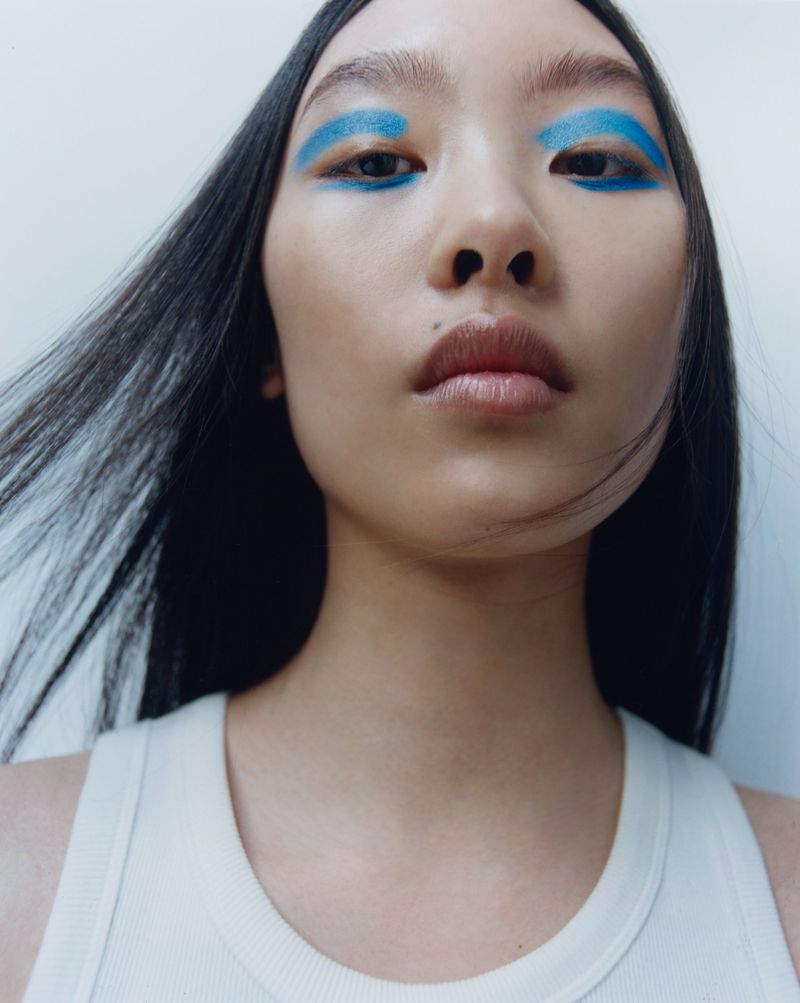 Close-up portrait of a model with vibrant blue eyeshadow and long dark hair against a plain white background.