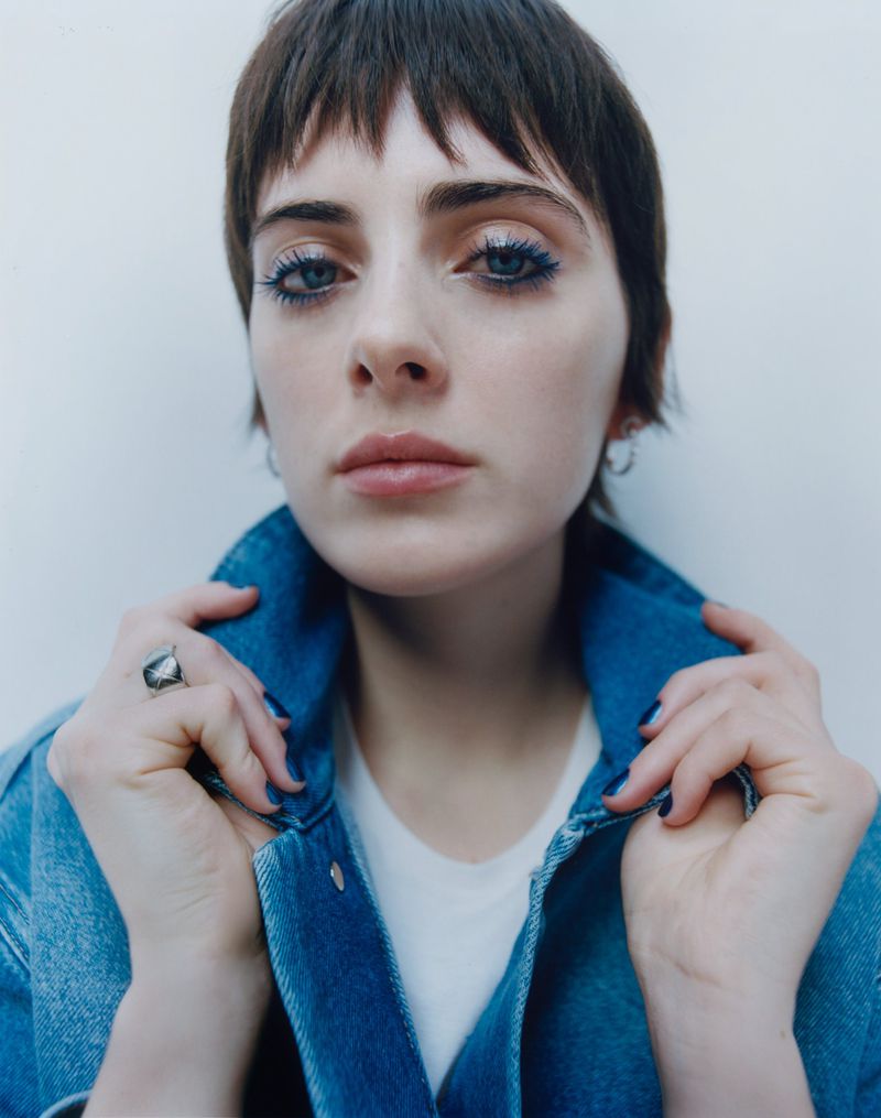Close-up portrait of a woman with dark short hair holding a blue denim jacket collar against a plain background