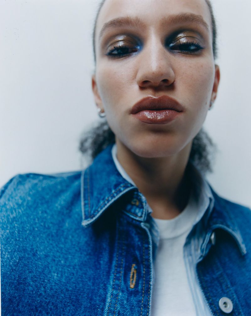 Close-up portrait of a model wearing a blue denim jacket and white t-shirt against a plain white studio background.