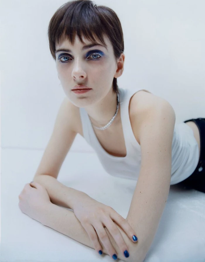 Model with blue eyeshadow and a white tank top leaning forward on a seamless white studio background