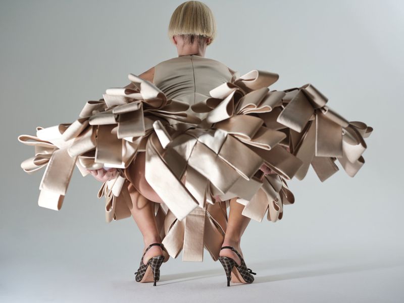 Model with blonde bob hair wearing an elaborate structured beige ribbon dress against a plain white studio backdrop.