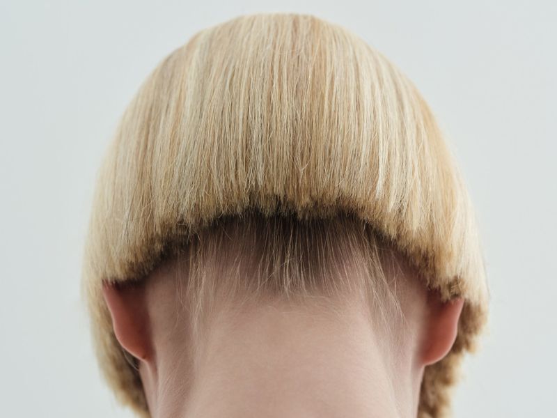 Rear view of a person with a short blonde bowl haircut showing the nape of the neck against a plain light background