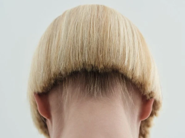 Rear view of a person with a short blonde bowl haircut showing the nape of the neck against a plain light background