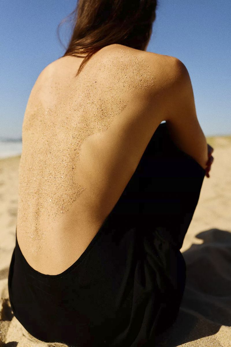 Rear view of a person wearing a black backless garment sitting on a sandy beach under a clear blue sky.