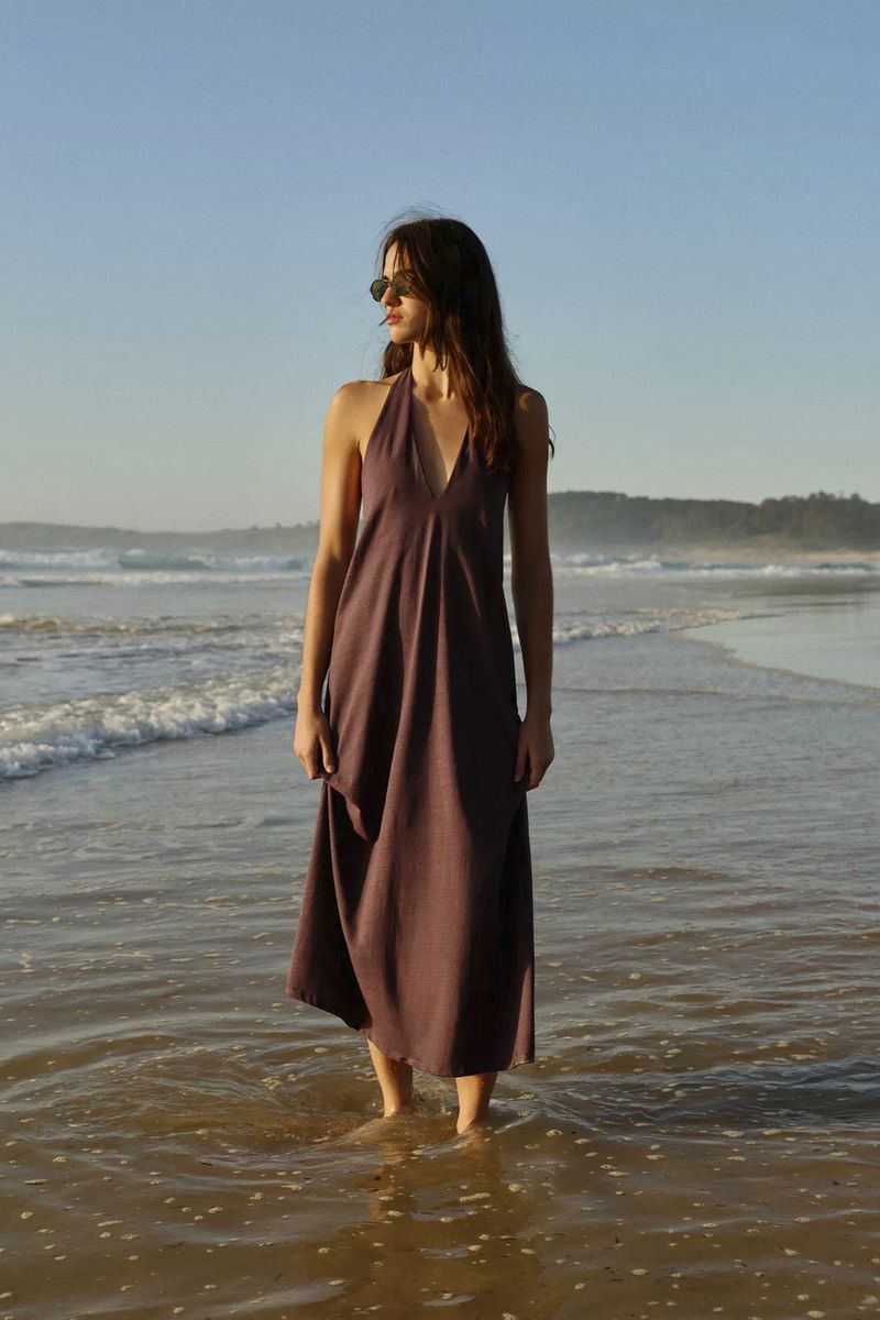 Woman wearing a halter neck dress standing ankle-deep in ocean surf on a sandy beach during sunset.