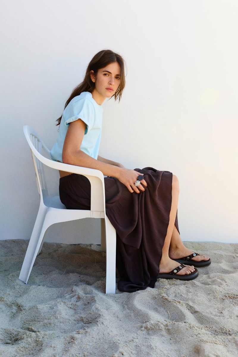 Woman seated on a white plastic chair on sand wearing a light blue t-shirt and dark skirt for Pull & Bear