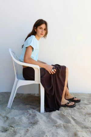 Woman seated on a white plastic chair on sand wearing a light blue t-shirt and dark skirt for Pull & Bear