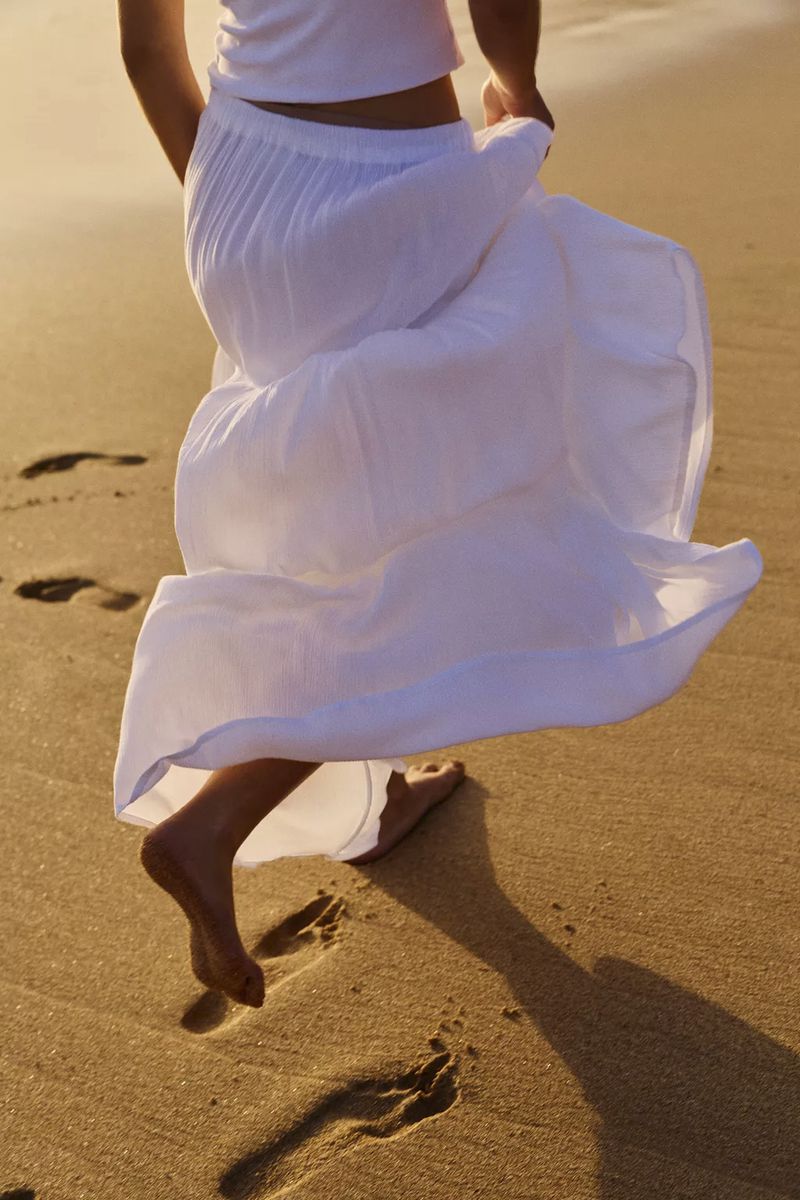 Woman in white dress runs barefoot on sandy beach leaving footprints at sunset. Lower back view of vacation freedom and wellness.