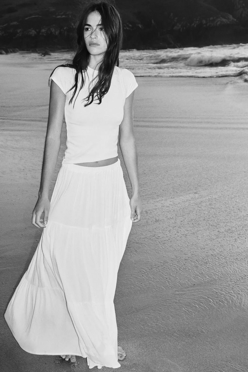Model in white t-shirt and maxi skirt standing on sandy beach in black and white fashion lookbook photo