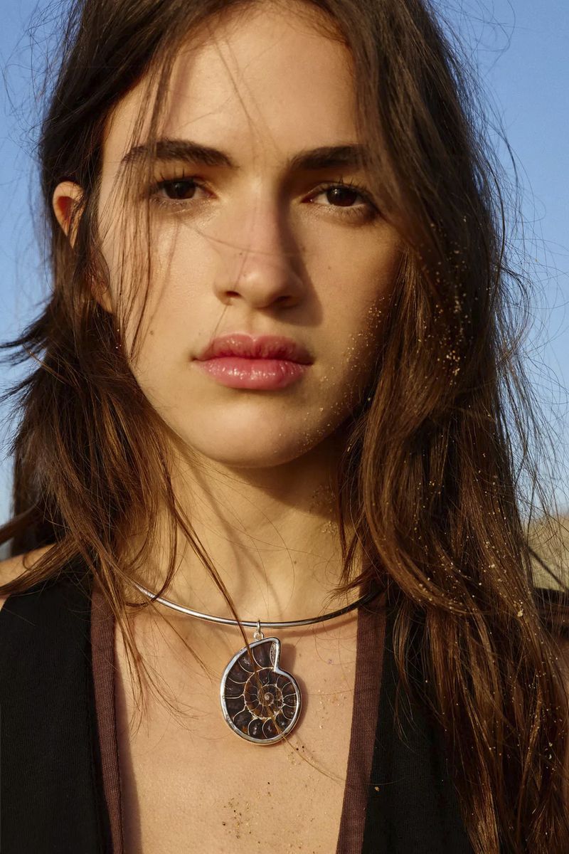 Close-up portrait of a young woman with long dark hair wearing a silver choker necklace with a large ammonite pendant against a clear sky.