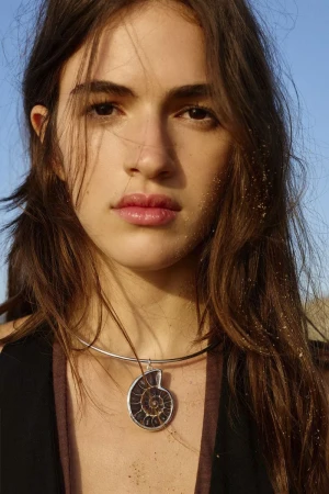 Close-up portrait of a young woman with long dark hair wearing a silver choker necklace with a large ammonite pendant against a clear sky.