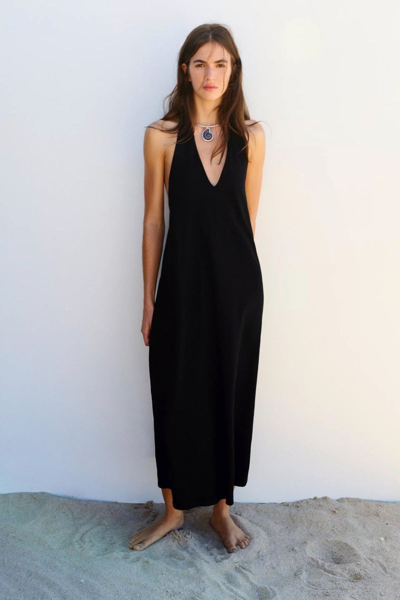 Model in a long black halter dress standing barefoot on sand against a plain white wall for Pull & Bear