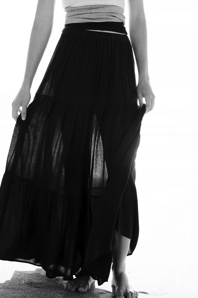 Model standing outdoors wearing a long black tiered skirt and layered top against a bright background