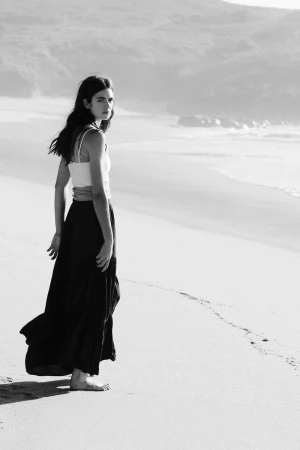Monochrome fashion photograph of a woman standing barefoot on a sandy beach looking back toward the camera.