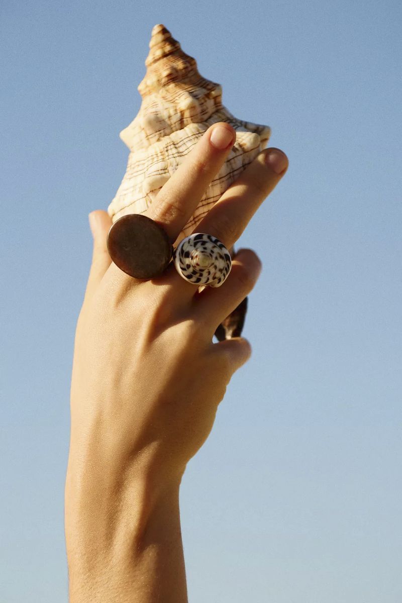 Hand holding a large spiral seashell against a clear blue sky, wearing two statement rings