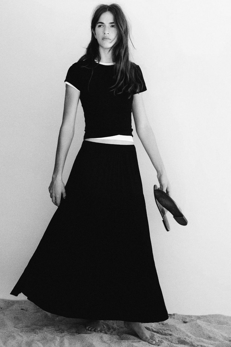 Model standing on sandy ground wearing a black t-shirt and long black skirt, holding sandals.