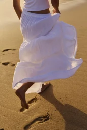 Model walking barefoot on a sandy beach wearing a flowing white tiered skirt and white top during golden hour sunlight.