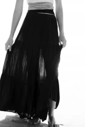 Model standing outdoors wearing a long black tiered skirt and layered top against a bright background