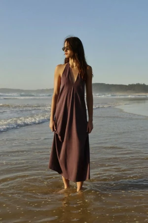 Woman wearing a halter neck dress standing ankle-deep in ocean surf on a sandy beach during sunset.