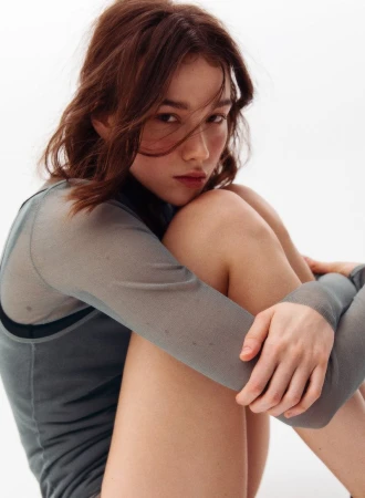 Young woman with reddish brown hair wearing a grey sheer long sleeve top hugging her knees against a plain white background