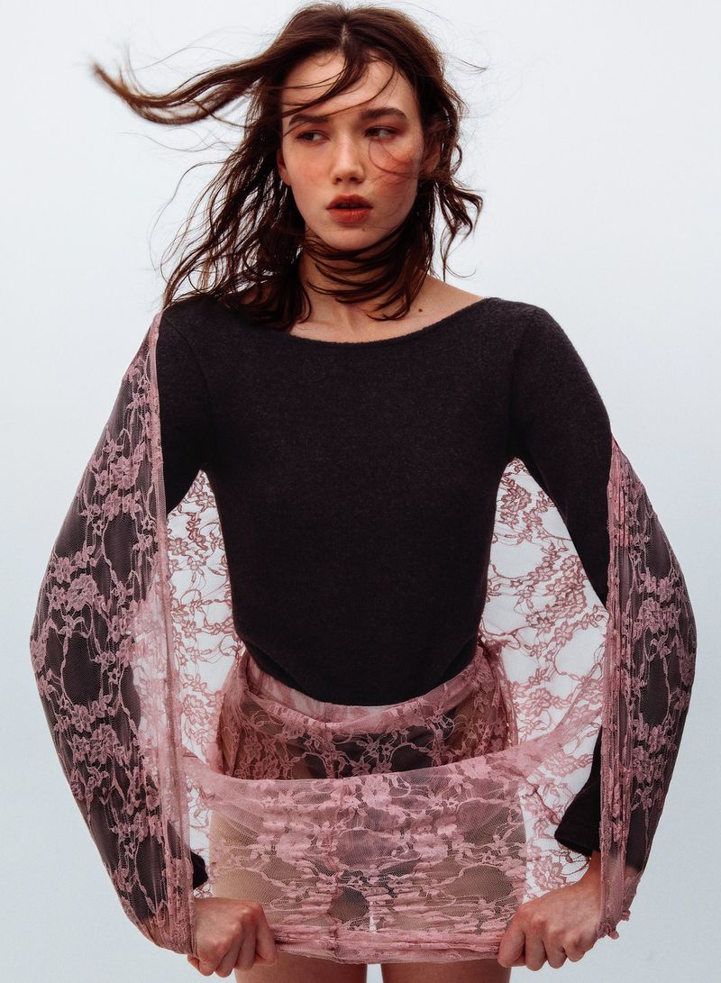 A model with long brown hair blowing in the wind wears a dark top and pink lace fabric against a white seamless background.